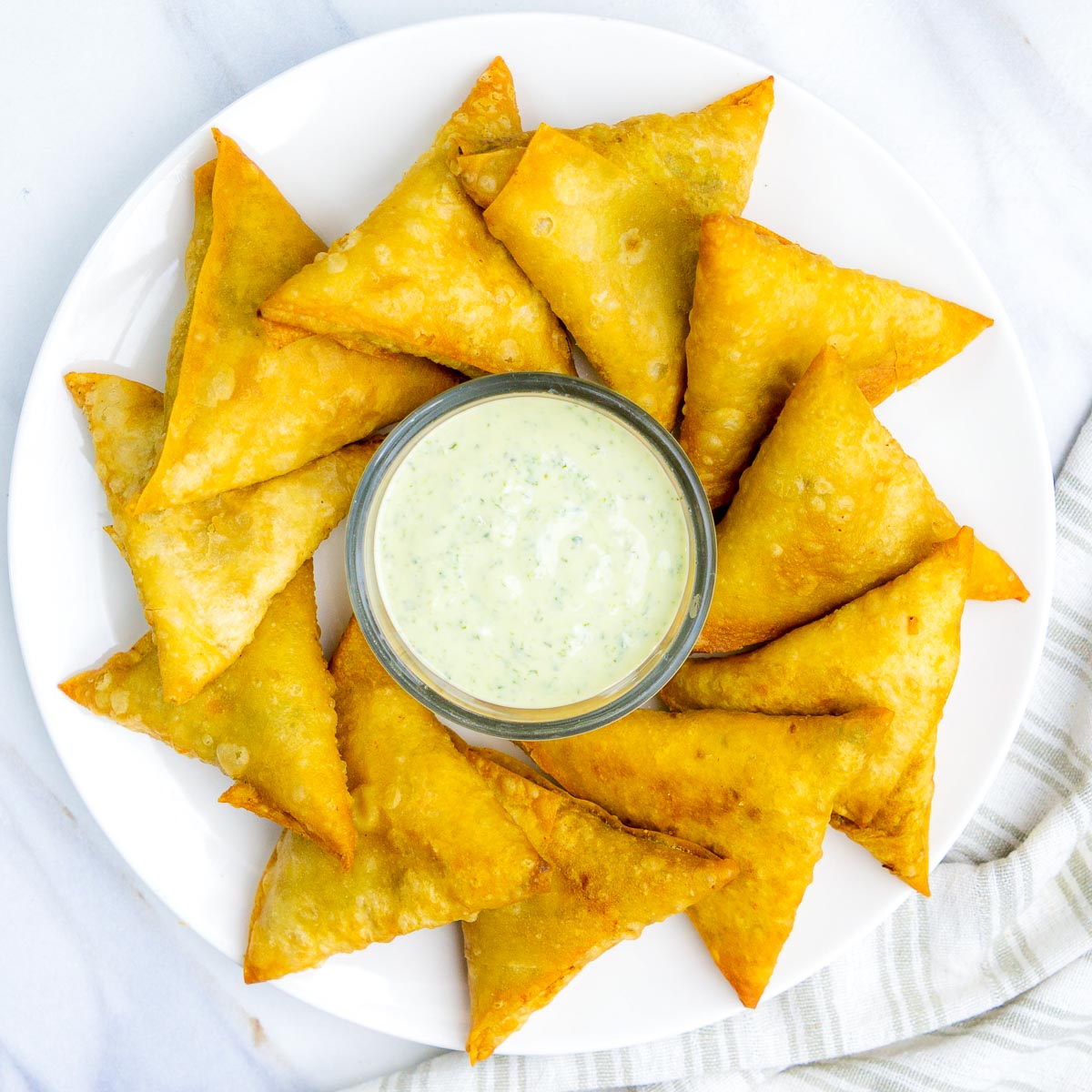 An overhead view of a circular white plate arranged with over a dozen golden-brown, triangular aloo samosas circling a small glass bowl of light green mint or cilantro chutney in the center. The plate sits on a white marble surface next to a striped linen napkin.