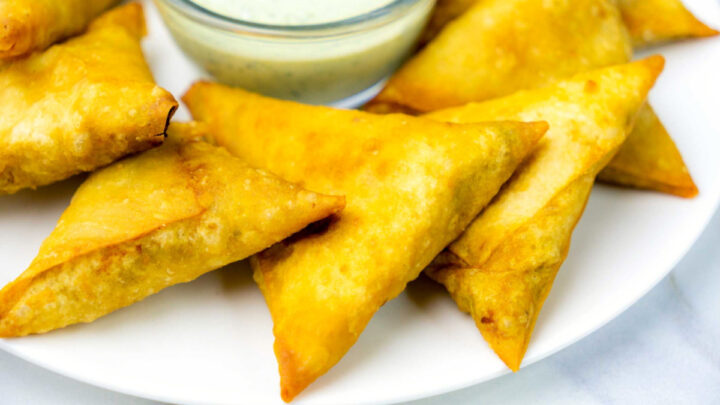 A close-up shot of golden-brown, crispy samosas piled on a white plate next to a small glass bowl of light green dipping sauce.
