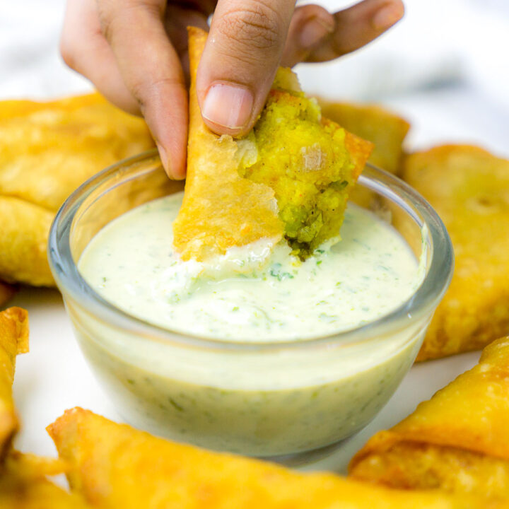 A close-up shot of a hand dipping a broken golden-brown samosa into a glass bowl of creamy light green chutney. The broken corner of the samosa reveals a textured yellow potato filling inside.