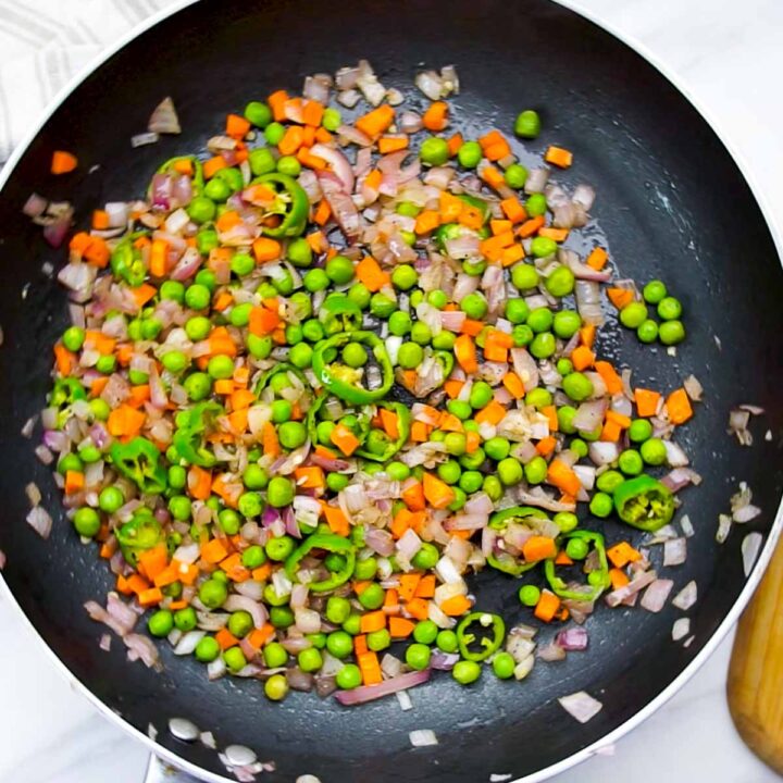 An overhead shot of a black frying pan containing a colorful mixture of sautéed vegetables, including green peas, diced orange carrots, chopped red onions, and sliced green chili peppers.