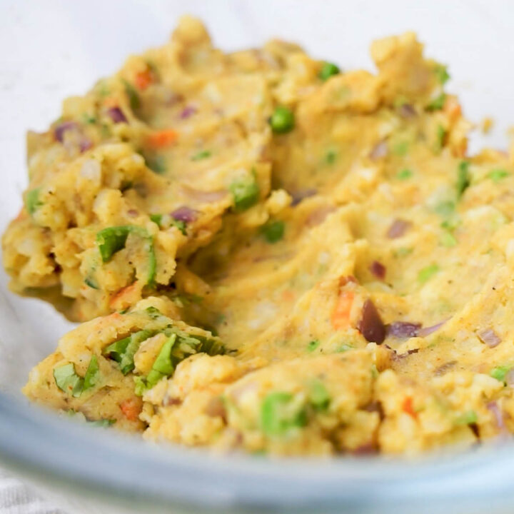 A close-up view of mashed yellow potatoes in a glass bowl, mixed with the sautéed peas, carrots, and onions to create a uniform samosa filling.