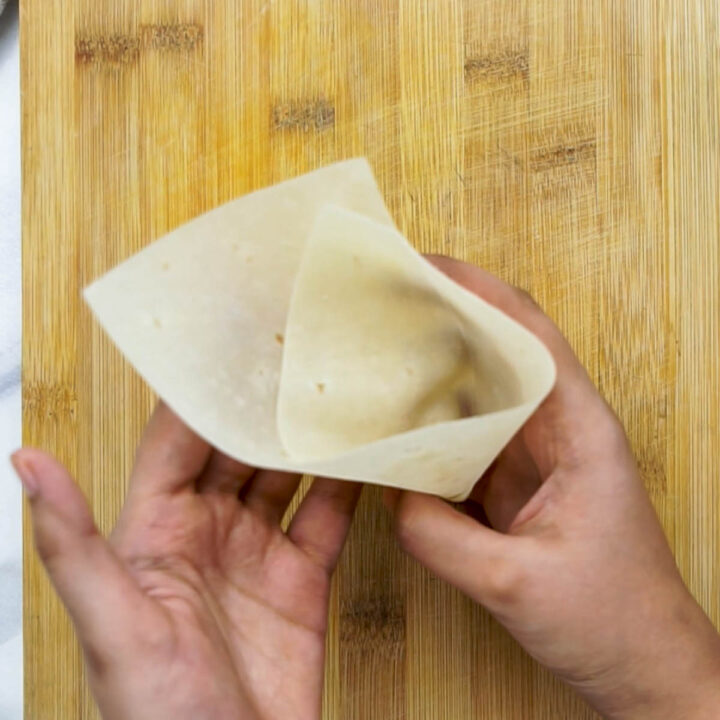An overhead shot of hands folding a thin, translucent pastry sheet into a cone shape over a wooden cutting board to prepare for the potato filling.