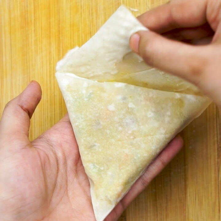 A close-up shot of hands folding the final corner of the thin pastry over the filled samosa to seal it into its triangular shape.