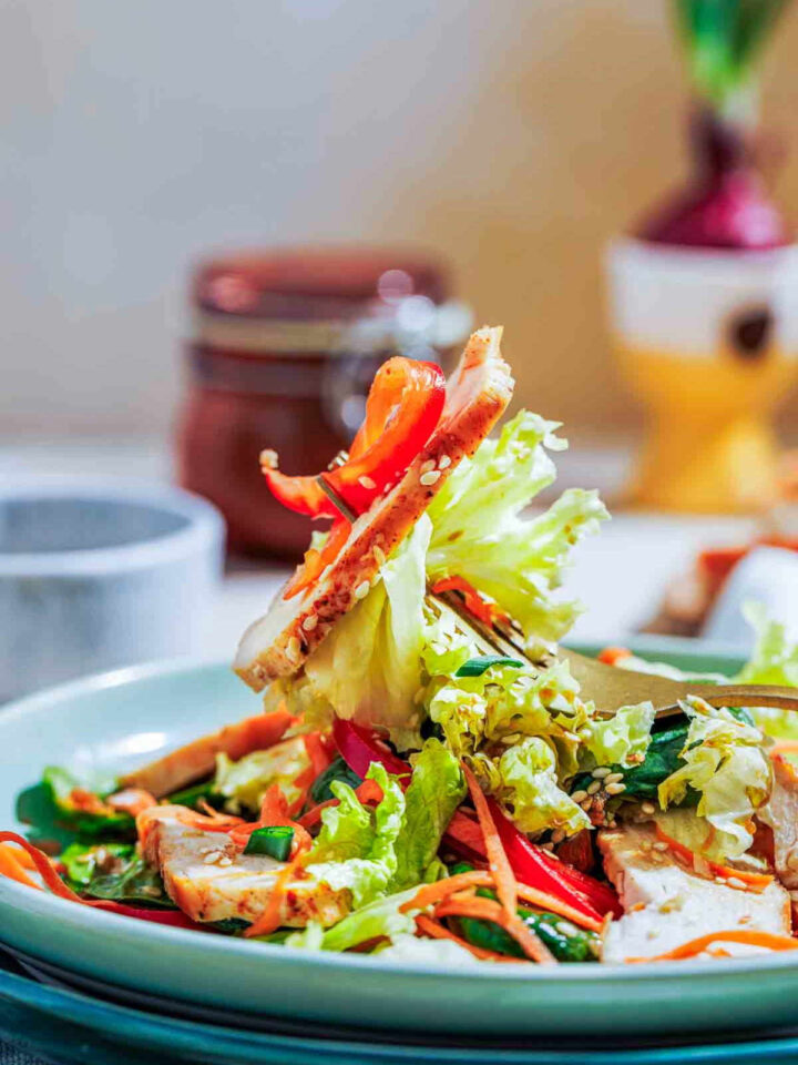 Close-up of a fork lifting a bite of chicken, lettuce, and red pepper from a sesame salad.
