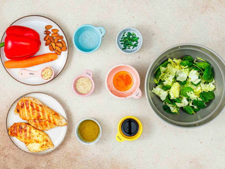Overhead view of ingredients including grilled chicken breasts, fresh greens, red pepper, carrots, almonds, sesame seeds, and various dressings in small bowls.