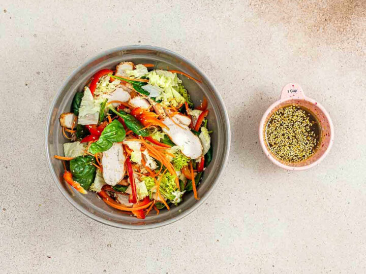 A large grey bowl filled with tossed chicken salad next to a small pink bowl of sesame dressing.