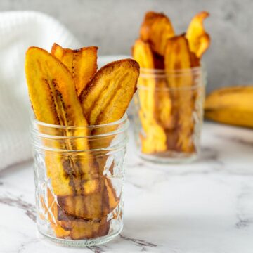 Crispy, golden-yellow banana or plantain chips (chifles) served upright in two glass mason jars on a marble surface.