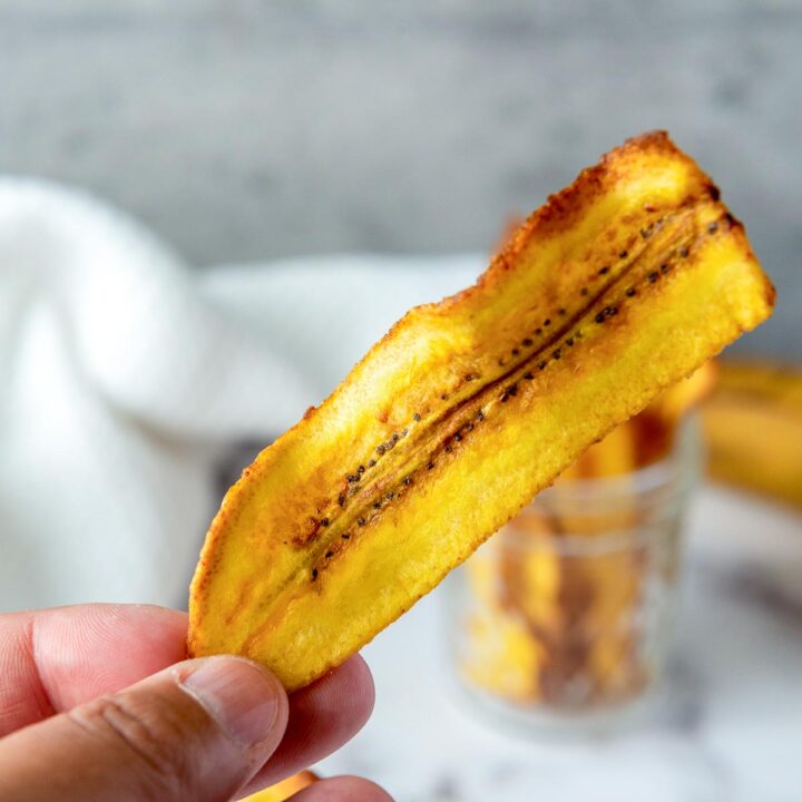 A hand holds up a single crispy, golden-yellow plantain chip (chifle) with a blurred glass jar in the background.