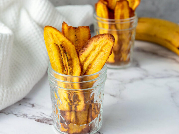 Crispy, golden-yellow plantain chips (chifles) served upright in two glass mason jars on a marble surface.