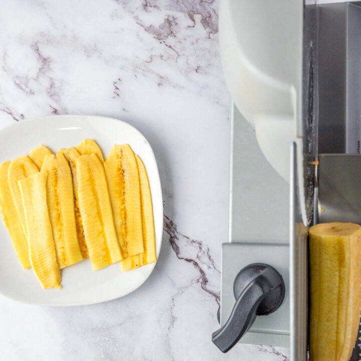 Long, thin slices of yellow plantain arranged on a white square plate next to a mandoline slicer on a marble counter.