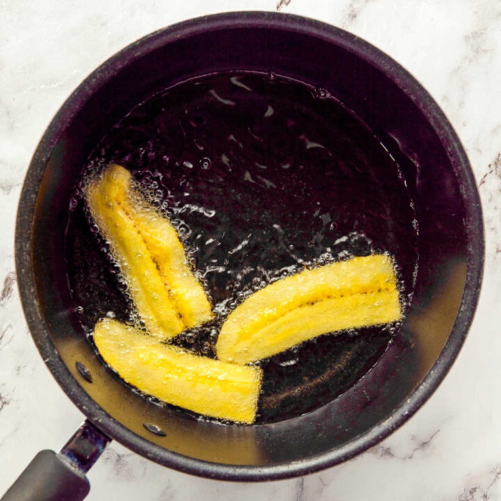 Three thick slices of yellow plantain frying in hot oil in a small black saucepan.