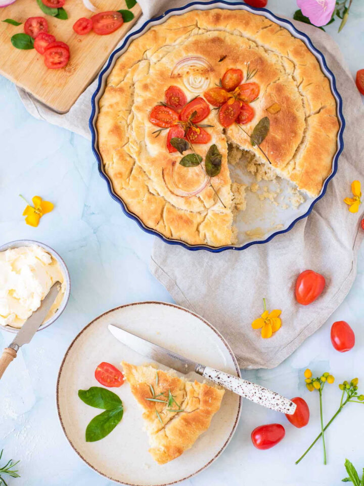 A large, round focaccia bread decorated with tomato "flowers" and onion rings in a blue baking dish, with a single slice served on a plate nearby.