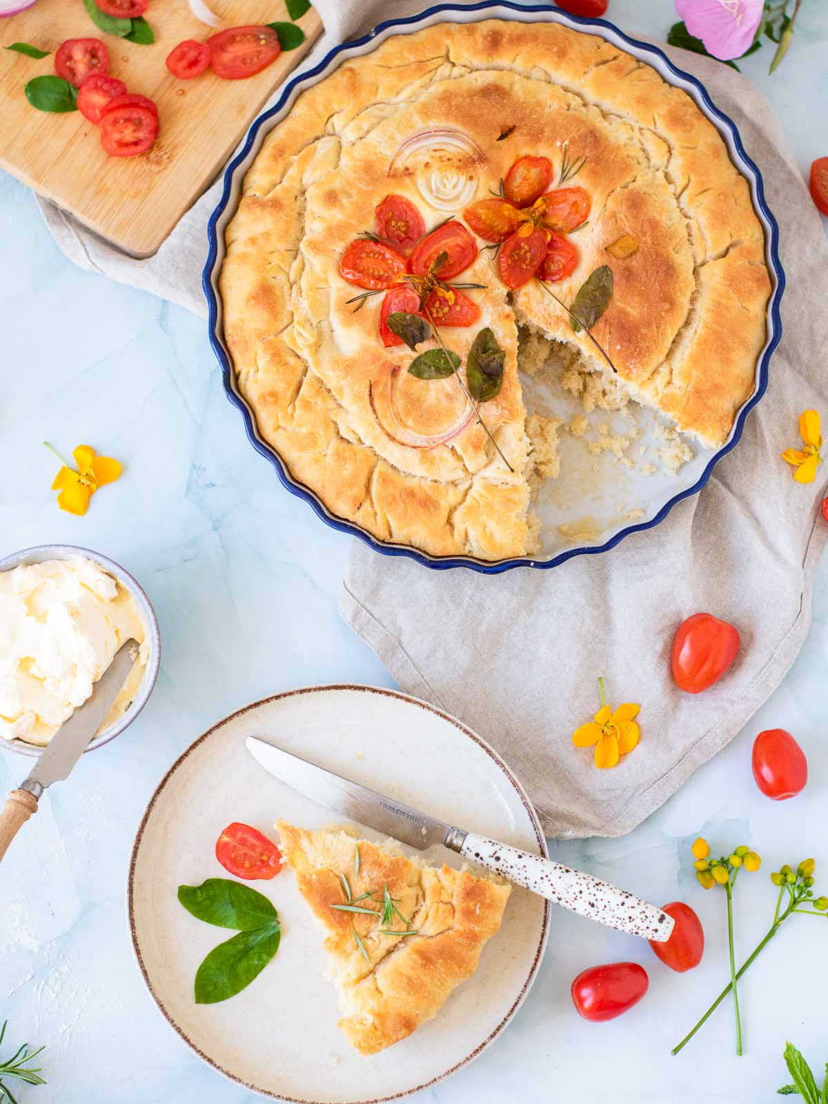 A large, round focaccia bread decorated with tomato "flowers" and onion rings in a blue baking dish, with a single slice served on a plate nearby.
