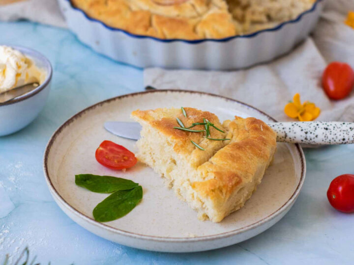 A top-down view shows the pale focaccia dough in a round blue dish, decorated with sliced cherry tomatoes and yellow edible flowers arranged to look like two blossoms with green leaf stems. Thin slices of red onion are placed at the top and bottom to resemble clouds or a sun.