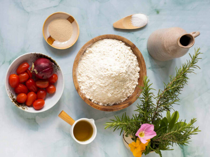 A top-down view of ingredients for focaccia, including a large wooden bowl of flour, small bowls of yeast, salt, olive oil, and cherry tomatoes, alongside fresh rosemary and edible flowers.