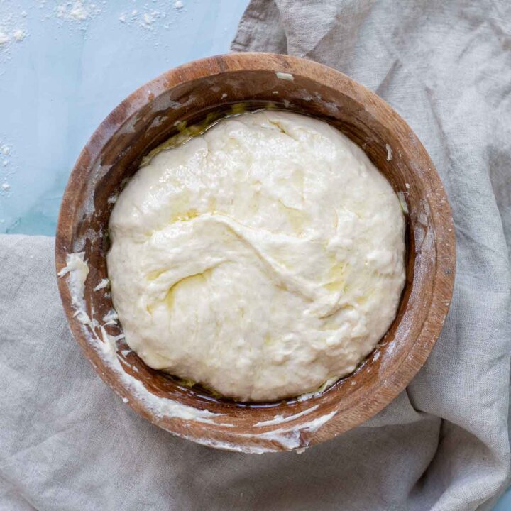 A top-down view of smooth, pale focaccia dough rising in a large wooden bowl, coated with a glistening layer of olive oil.