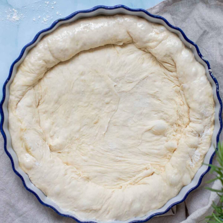 A top-down view of pale focaccia dough pressed into a round, blue fluted baking dish, showing a thick, puffy outer rim.