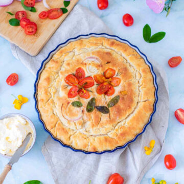 A top-down view shows the golden-brown baked focaccia in its blue dish. The tomato flowers and onion slices have roasted into the surface, which appears crisp and textured. A bowl of whipped butter and scattered fresh tomatoes surround the dish on a light blue background.