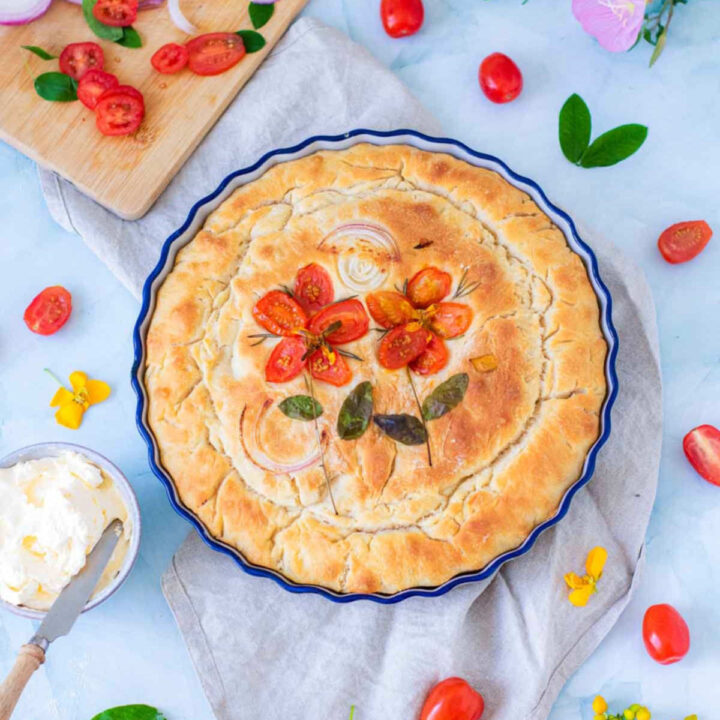 A top-down view shows the golden-brown baked focaccia in its blue dish. The tomato flowers and onion slices have roasted into the surface, which appears crisp and textured. A bowl of whipped butter and scattered fresh tomatoes surround the dish on a light blue background.