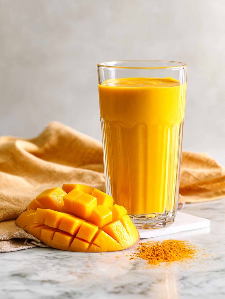 Mango turmeric smoothie in tall glass with mango cheekon the side.
