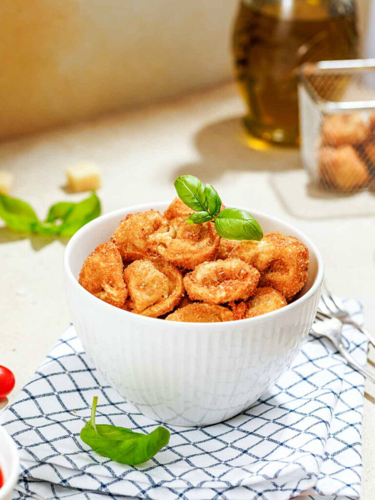 A white bowl filled with golden-brown parmesan fried tortellini garnished with fresh basil leaves.