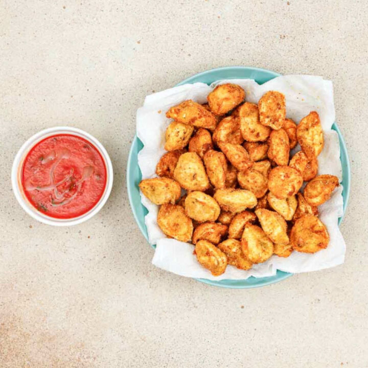 Fried tortellini draining on a paper towel-lined blue plate next to a bowl of marinara.