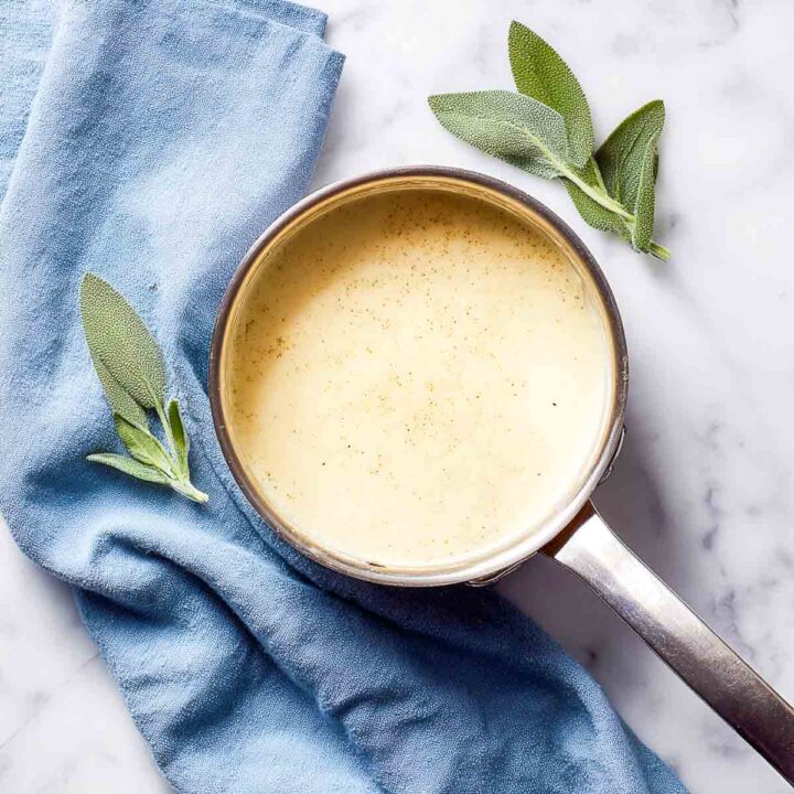 Creamy cheese sauce simmering in a stainless saucepan with fresh sage nearby.