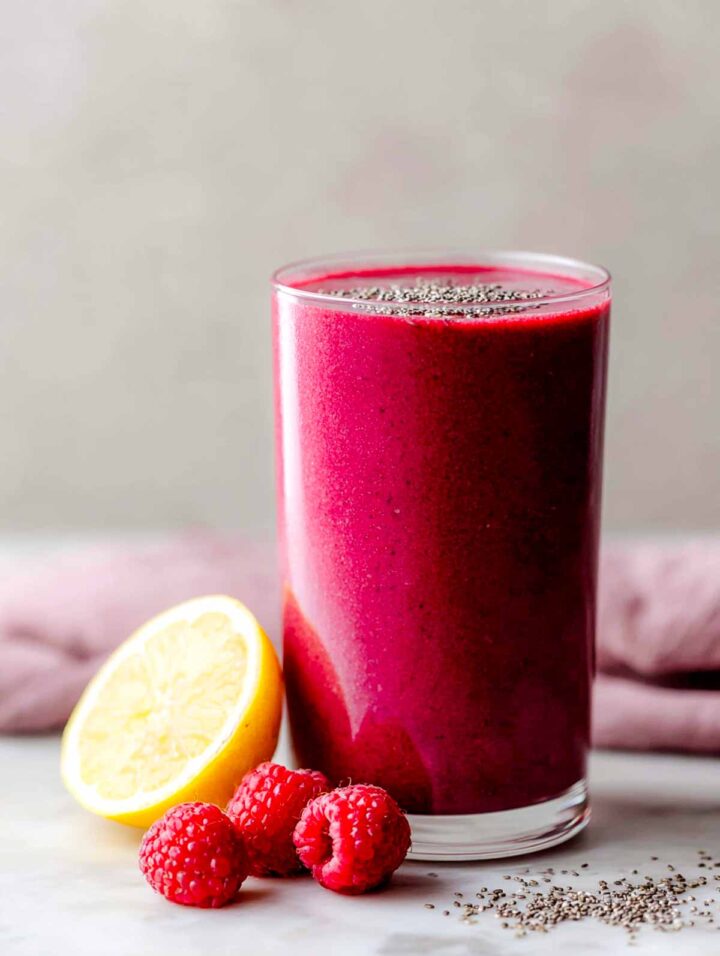 Raspberry beet smoothie in tall glass with lemon and raspberries placed next to the glass.