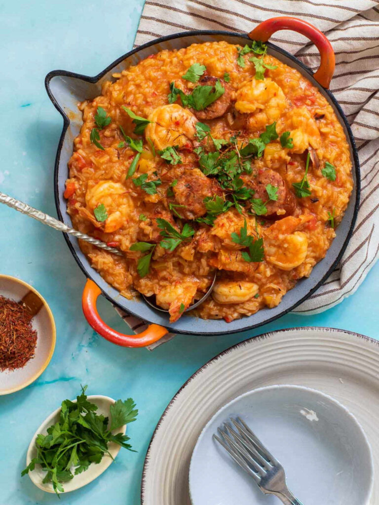 An overhead shot of a skillet filled with vibrant saffron paella, featuring shrimp and sliced sausage, garnished with fresh parsley and saffron threads, surrounded by a small bowl of saffron, fresh herbs, and dinner plates.