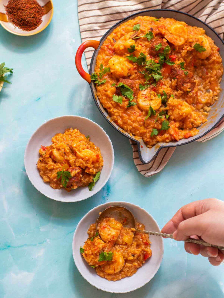 A small white bowl of saffron shrimp paella, garnished with parsley, sits in the foreground. The paella pan is in the background.