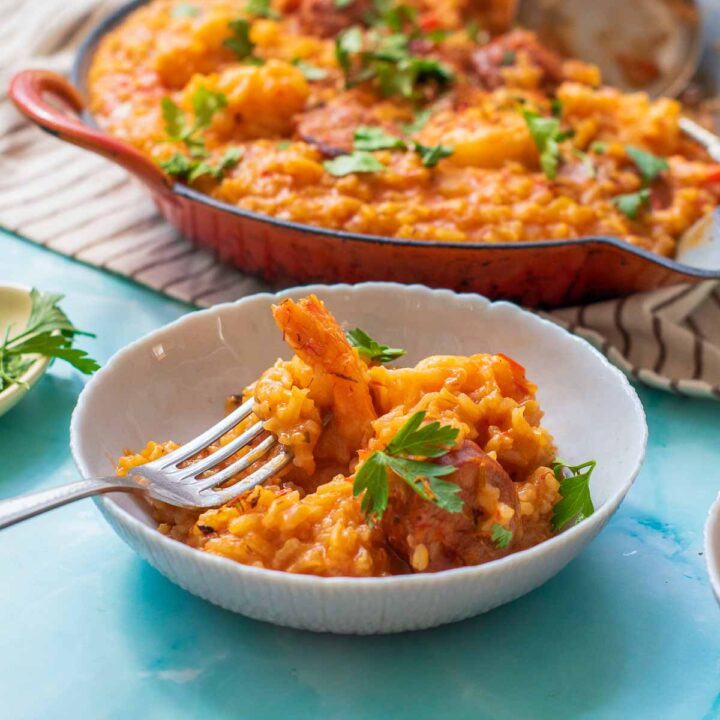 An overhead shot of a skillet filled with vibrant saffron paella, featuring shrimp and sliced sausage, garnished with fresh parsley and saffron threads, surrounded by a small bowl of saffron, fresh herbs, and dinner plates.
