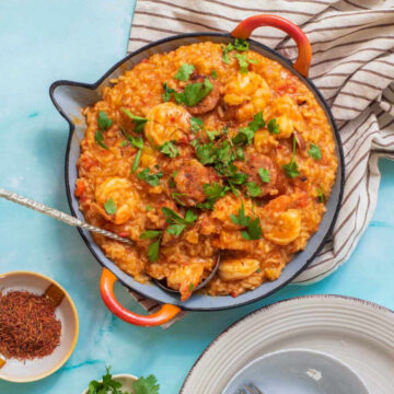 An overhead shot of a skillet filled with saffron paella, with shrimp and sliced sausage, garnished with fresh parsley and saffron threads, surrounded by a small bowl of saffron, fresh herbs, and dinner plates.