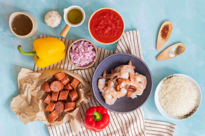 An overhead flat-lay shot of paella ingredients arranged on a blue surface, including raw shrimp, sliced chorizo, chopped onions, bell peppers, white rice, broth, saffron threads, and crushed tomatoes.