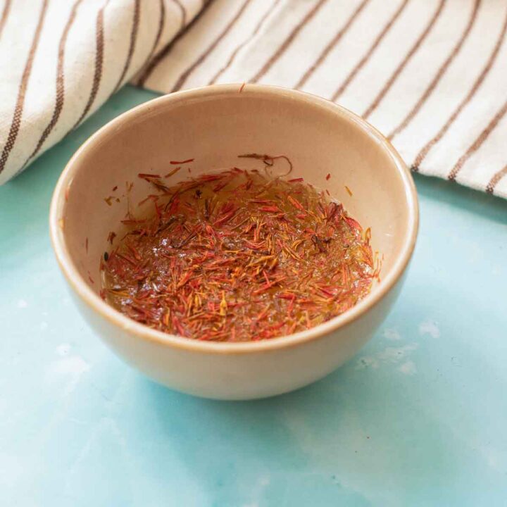 A small beige bowl containing saffron threads soaking in liquid, resting on a blue surface next to a striped kitchen towel.