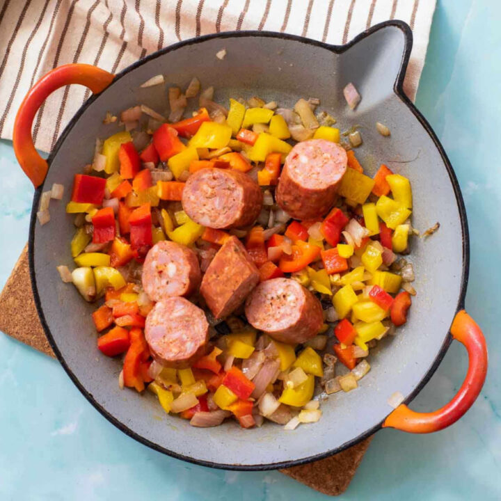 Sliced chorizo sausage cooking with diced red and yellow bell peppers and onions in a paella pan.