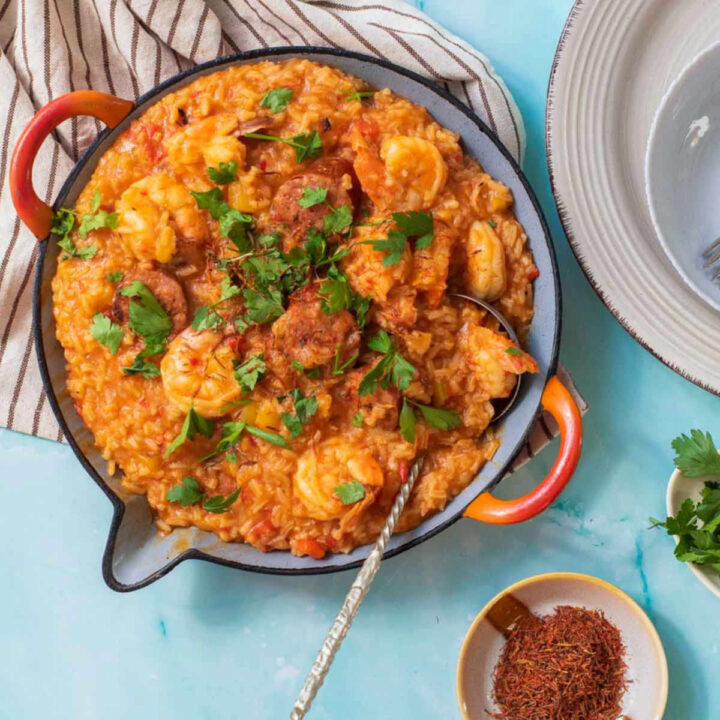 A completed paella dish in a gray pan with orange handles, featuring saffron-colored rice, shrimp, slices of chorizo, and fresh parsley garnish. A serving spoon is resting in the dish.