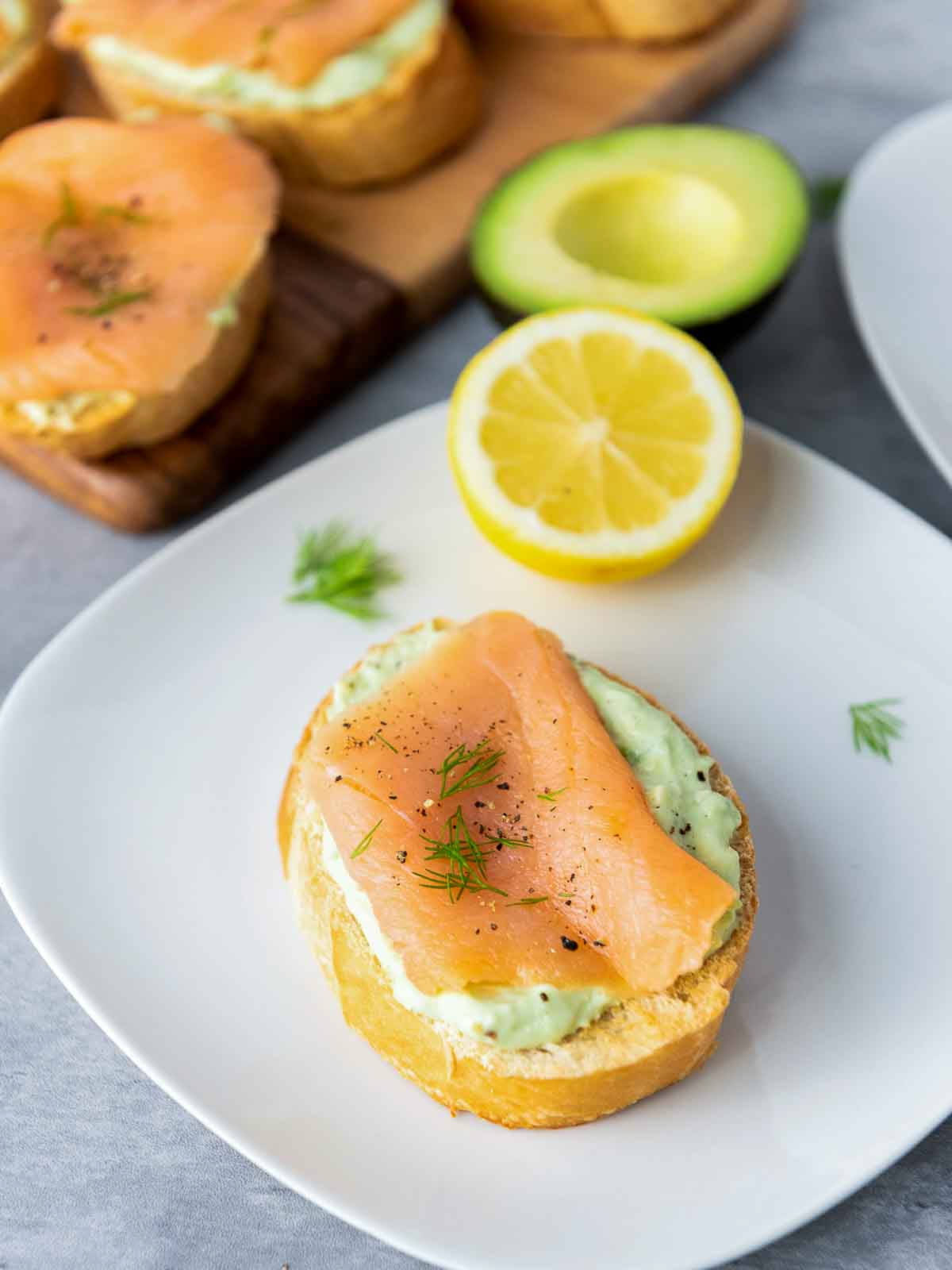 A piece of smoked salmon and avocado bruschetta on a white plate, topped with fresh dill and black pepper, with a lemon half and avocado in the background.