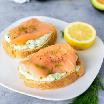 Two slices of smoked salmon and avocado bruschetta on a white plate, seasoned with black pepper and fresh dill, served with a lemon half.