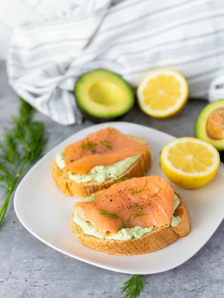 Two completed bruschettas served on a white plate, accompanied by a fresh lemon half and fresh dill, with avocados and citrus visible in the background.