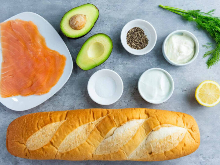 Two slices of crusty bread topped with creamy avocado spread and smoked salmon, garnished with fresh dill and cracked pepper, with a lemon half nearby.
