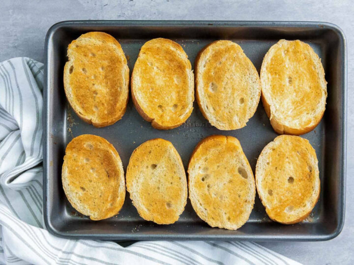 Eight oval slices of bread arranged on a dark baking sheet, toasted until golden brown.