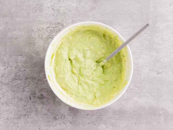 A white bowl containing a pale green, whipped mixture, likely the avocado combined with the creamy white spreads, being stirred with a metal spoon.
