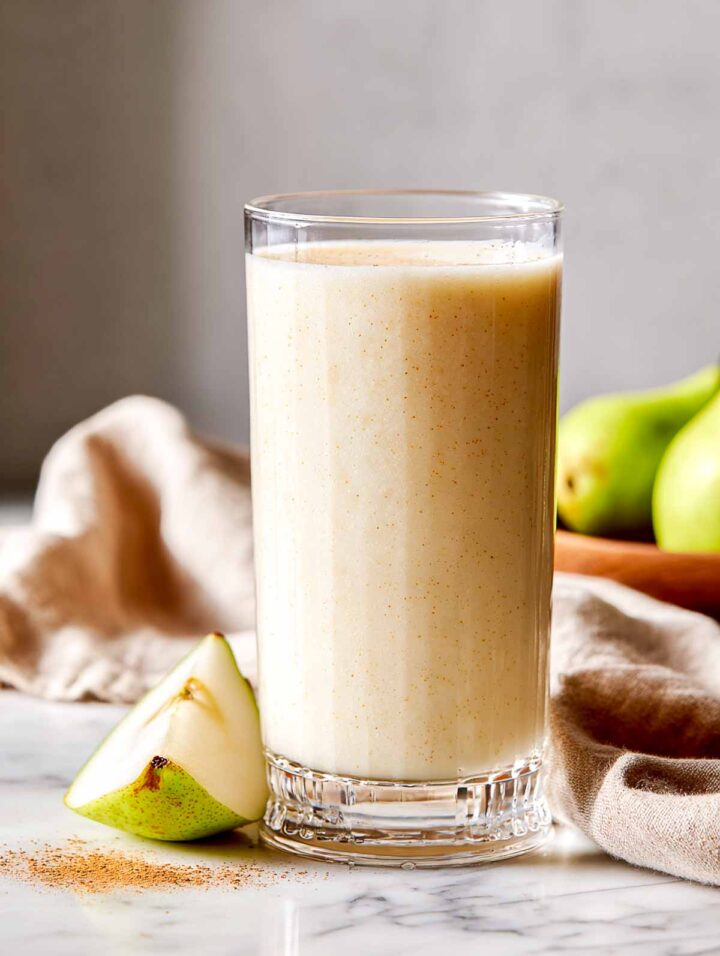 Pear cinnamon smoothie in tall glass with pear slice next to it.
