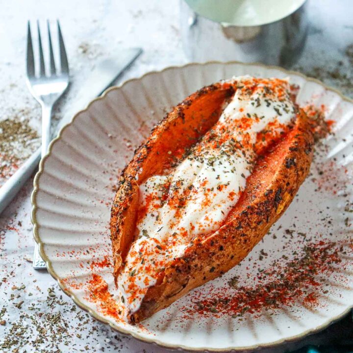 A roasted sweet potato is split down the center on a fluted white plate, filled with creamy white Greek yogurt and topped with more red spices and herbs.