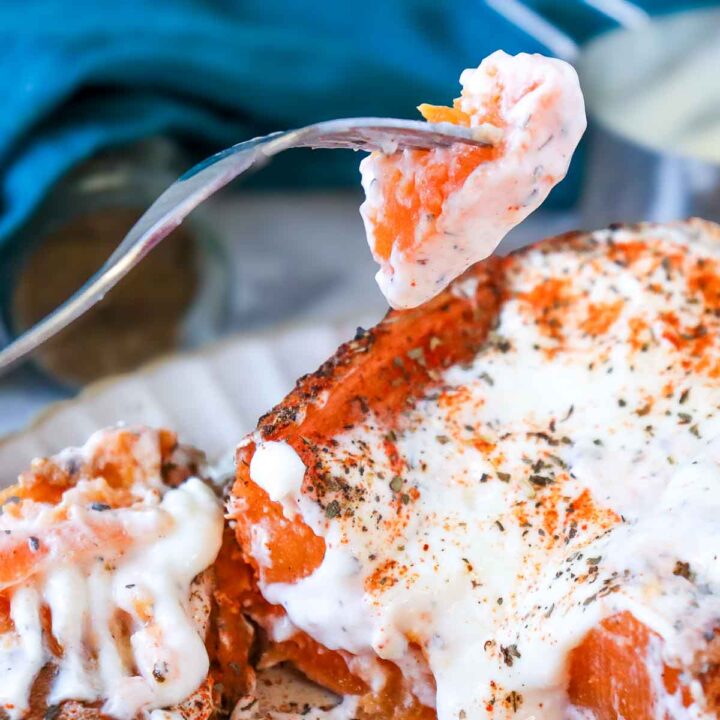 A fork lifts a bite-sized piece of orange sweet potato coated in creamy white Greek yogurt. Below, the main sweet potato is topped with yogurt and a heavy dusting of red paprika and dried green herbs.