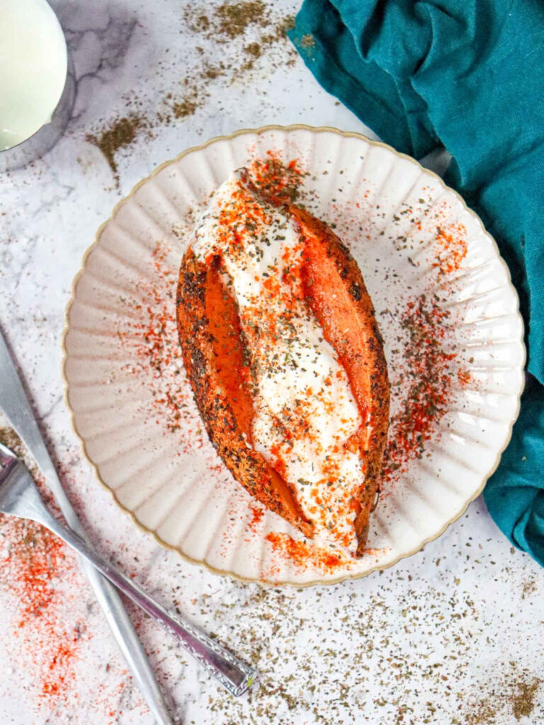 A top-down view of a whole roasted sweet potato split open on a white scalloped plate, filled with thick white Greek yogurt and heavily dusted with red paprika and dried herbs.