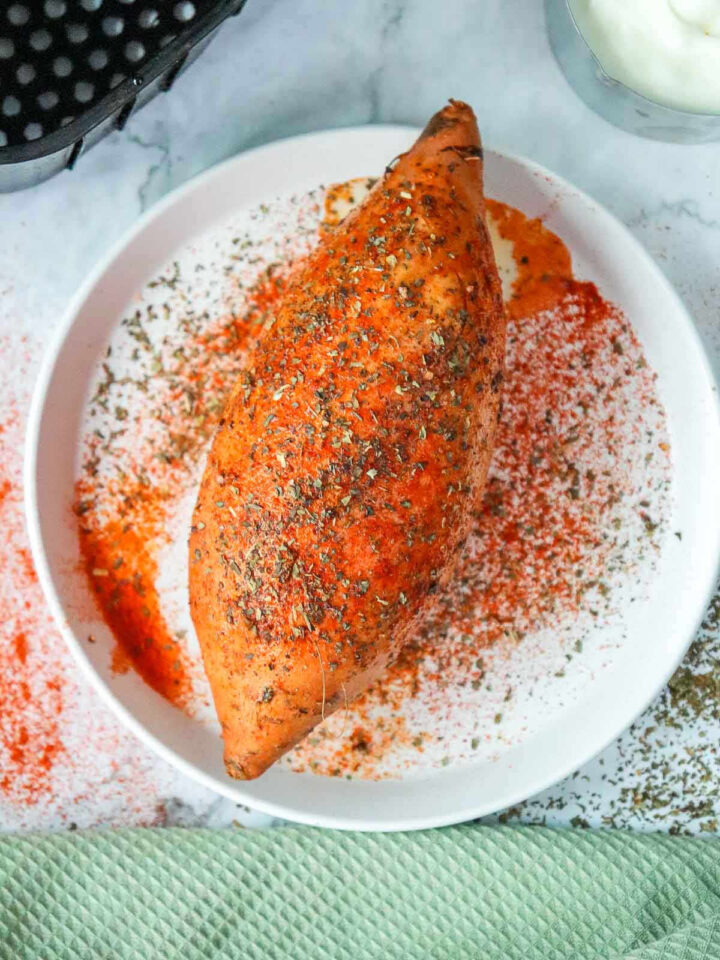 A whole sweet potato on a plate, heavily coated in red spices and dried herbs.