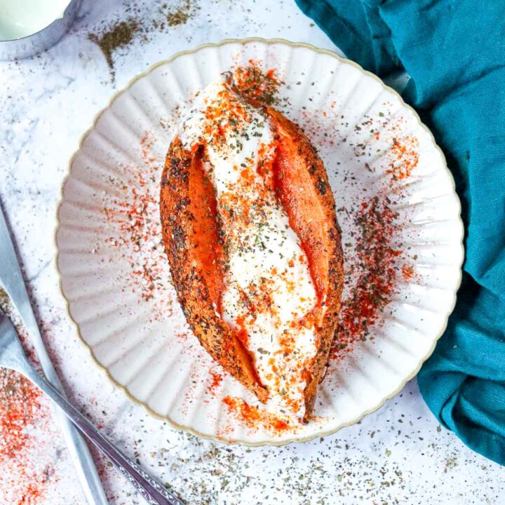 A top-down view of a roasted sweet potato split open on a white scalloped plate, filled with creamy white Greek yogurt and heavily dusted with red paprika and dried green herbs.
