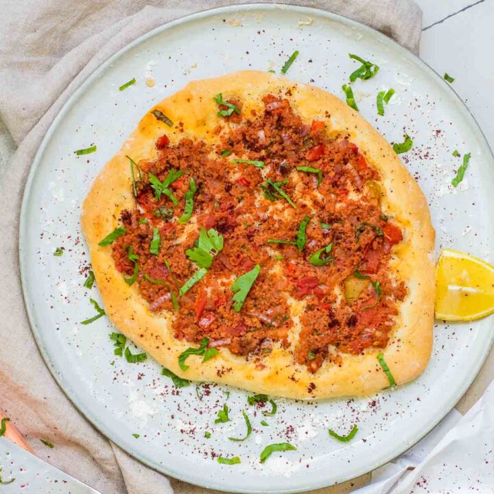 A close-up shot of a round, thin Turkish lahmacun flatbread on a light-colored plate, topped with a savory layer of minced meat, tomatoes, and onions. The dish is garnished with fresh green parsley and a dusting of red sumac, accompanied by lemon wedges and a small bowl of sumac in the background.
