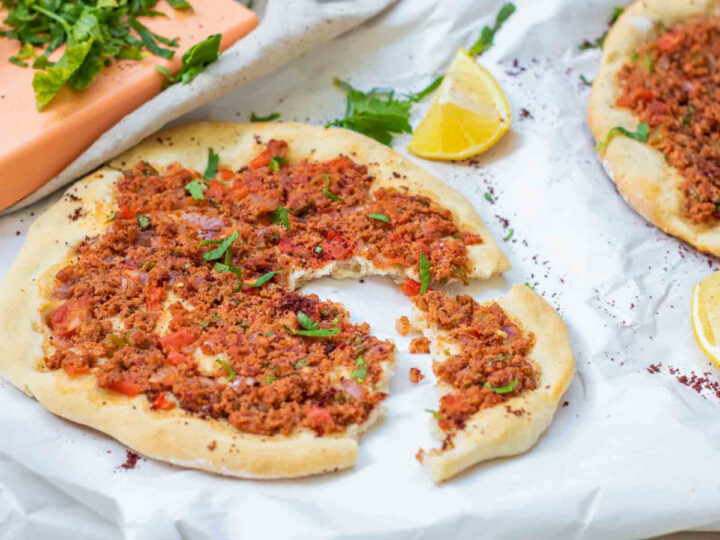 A close-up of a baked lahmacun on white paper with a piece torn away, showing the thin crust and savory topping, surrounded by fresh herbs, lemon wedges, and scattered sumac.